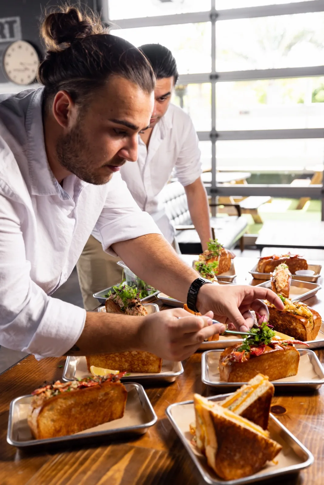 Two Lobster Lab chefs plating lobster rolls and grilled cheese onto steel trays lined up along a wooden table